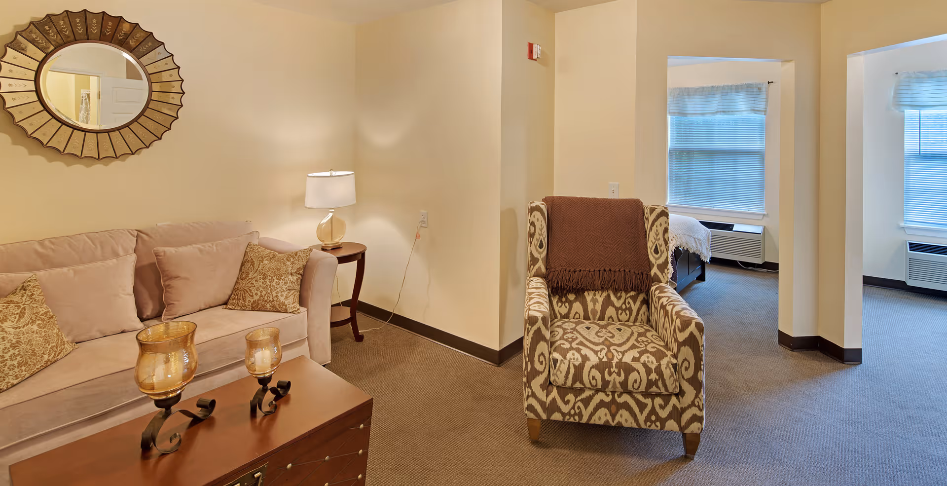 A cozy living area in a senior living facility featuring a beige sofa with patterned cushions, a decorative round mirror on the wall, a side table with a lamp, and a patterned armchair with a brown throw blanket. In the background, two windows with blinds and a glimpse of a bedroom with a bed are visible.