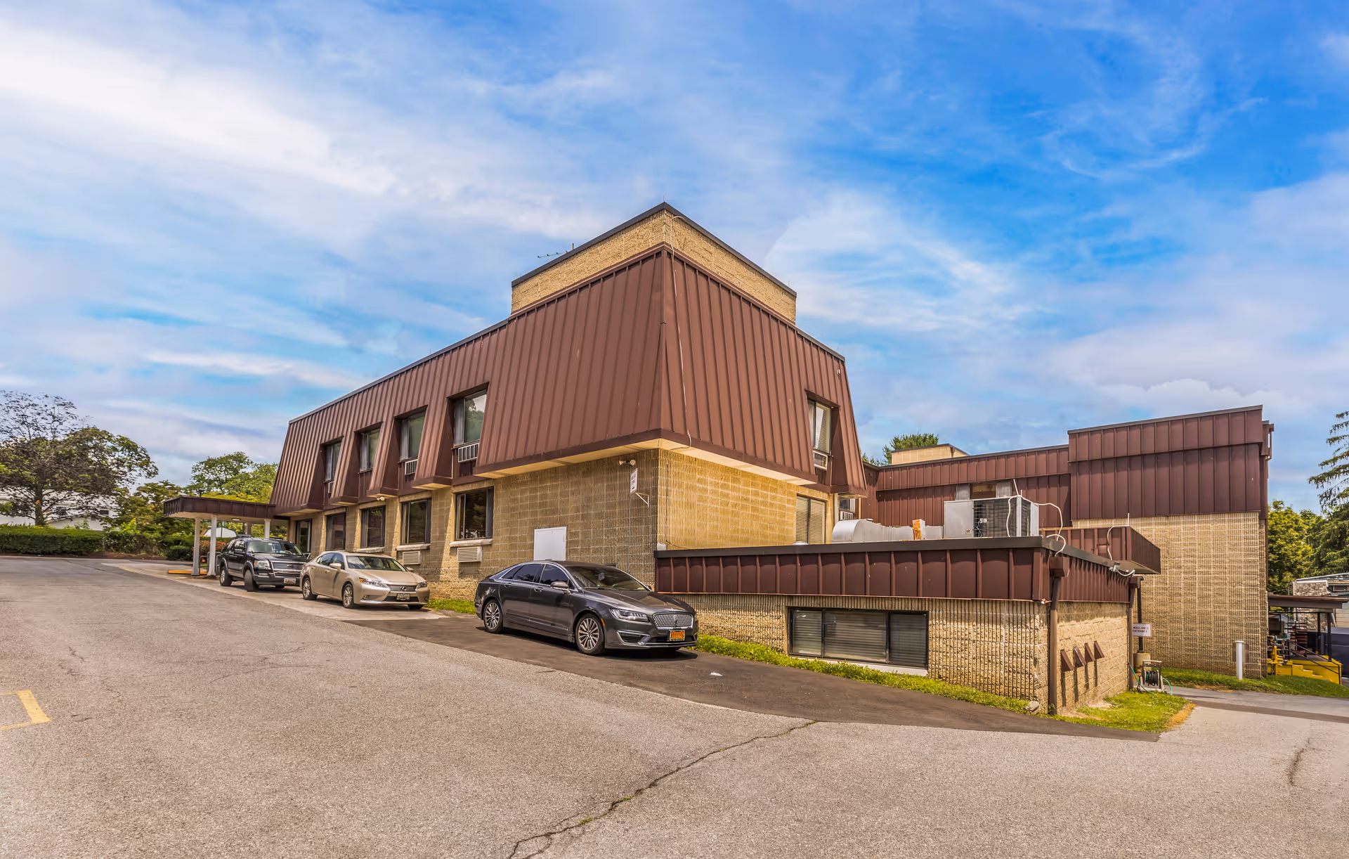 Exterior view of a two-story healthcare facility building with brown metal siding and beige brick walls. Several cars are parked along the driveway in front of the building under a partly cloudy blue sky.