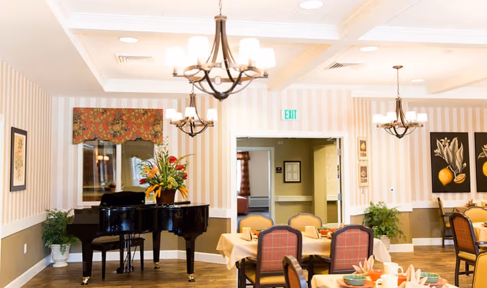 A dining room with several tables set for a meal, featuring chairs with red plaid upholstery. A black grand piano with a floral arrangement on top is positioned against a wall with striped wallpaper. The room is lit by multiple chandeliers and decorated with framed artwork and plants.