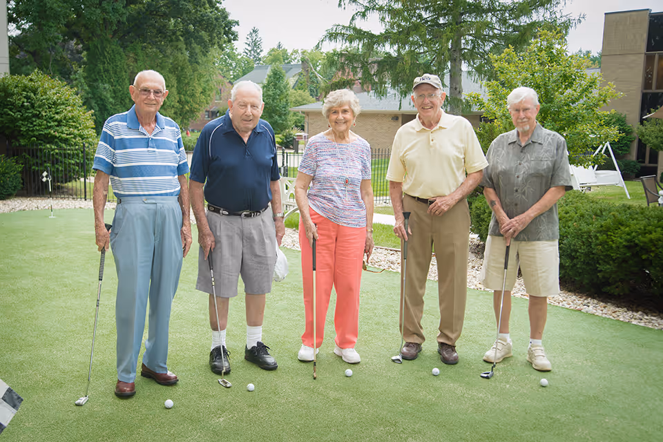 Five elderly individuals standing on a putting green holding golf clubs, smiling at the camera. They are outdoors with trees, bushes, and buildings in the background.