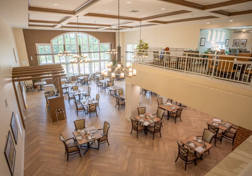 A spacious dining room in Juniper Village at Hamilton with multiple tables and chairs arranged neatly. The room features large windows allowing natural light to fill the space, elegant chandeliers hanging from a coffered ceiling, and a mezzanine level with additional seating where a few people are seated.