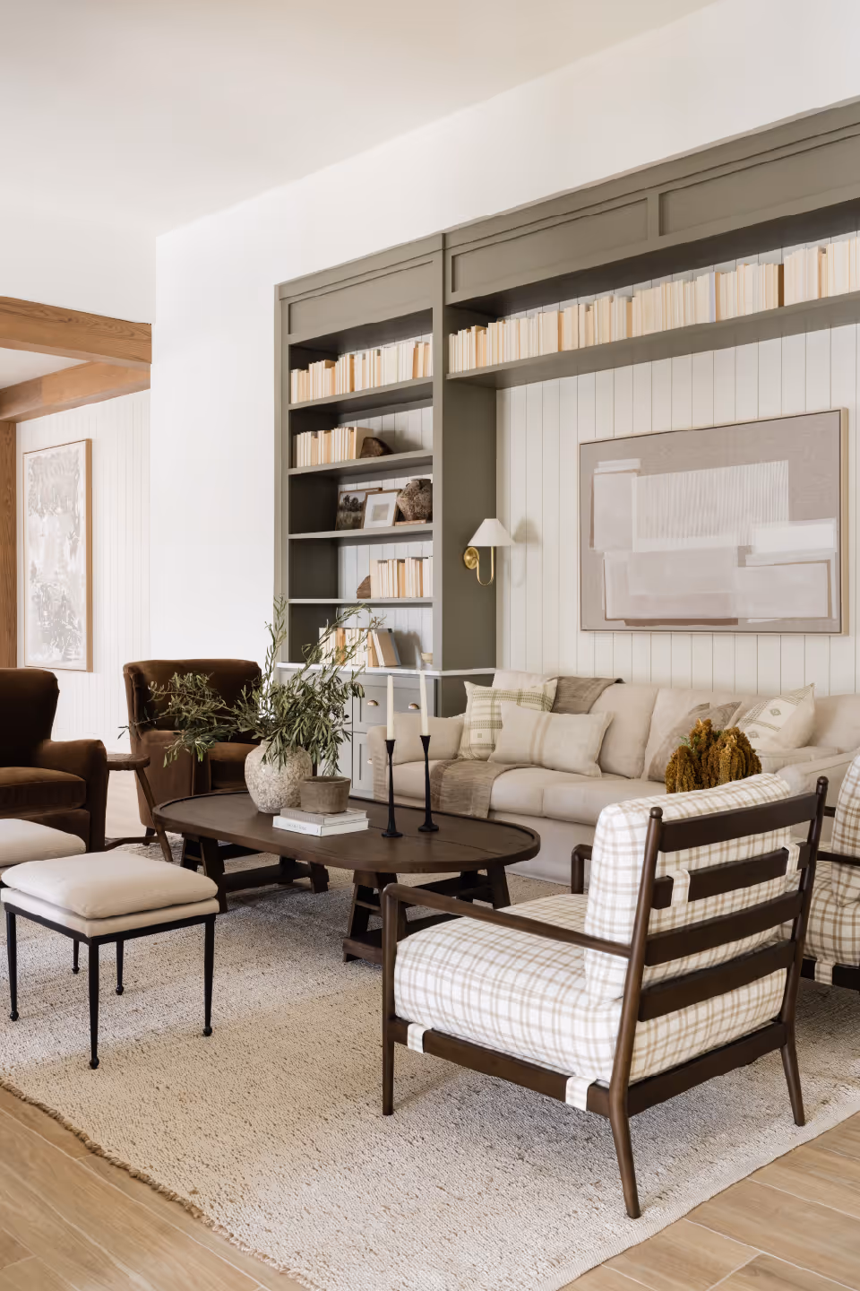 A cozy living room with a beige sofa, two plaid armchairs, and two brown armchairs arranged around a dark wooden coffee table. The coffee table holds a vase with greenery, two candlesticks, and some books. Behind the sofa is a built-in bookshelf painted in olive green, filled with books and decorative items. The walls are light-colored with vertical paneling, and there is a large abstract painting above the sofa. The floor is wooden with a light textured rug covering the seating area.