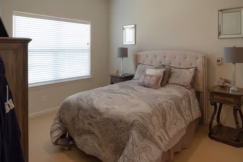 A cozy bedroom with a large window covered by white blinds on the left wall. The room features a bed with a beige patterned comforter and multiple pillows, flanked by two wooden nightstands each holding a lamp. The walls are light-colored, and there are two small framed mirrors above the nightstands.