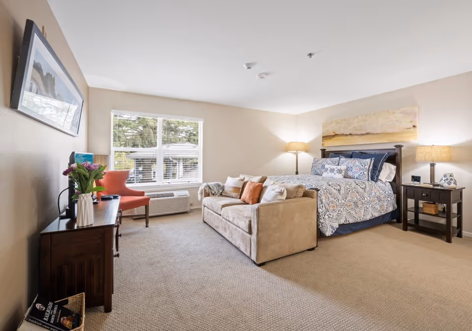 A well-lit bedroom with a large bed adorned with patterned bedding and multiple pillows. In front of the bed is a beige sofa with several cushions. To the right of the bed is a dark wooden nightstand with a lamp and decorative items. On the left side of the room, there is a wooden dresser with a vase of flowers and a TV mounted above it. A coral-colored chair is positioned near a window with white blinds, and an air conditioning unit is installed below the window. The walls are painted light beige, and the floor is carpeted.