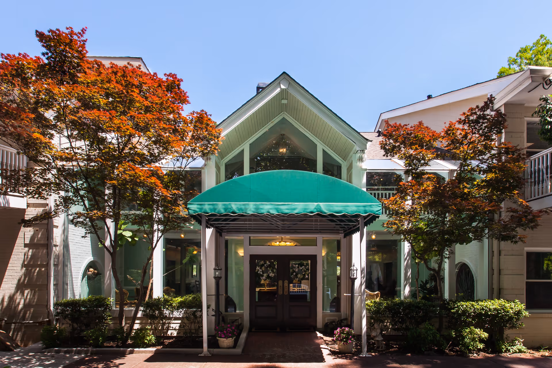 Front entrance of a senior living facility with a green awning above double doors, flanked by large windows and decorative plants. Two trees with red and orange leaves frame the entrance under a clear blue sky.