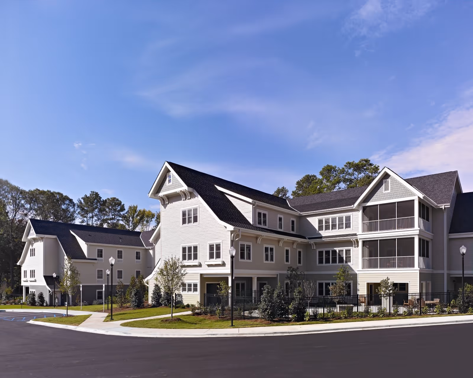 Exterior view of a large, multi-story residential building with light-colored siding and dark roofing, surrounded by landscaped greenery and a paved driveway under a clear blue sky.