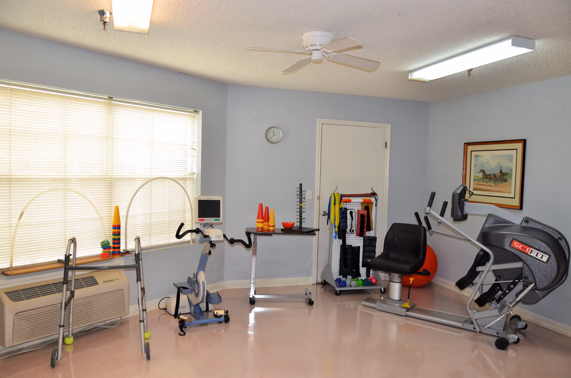 A small exercise room with light blue walls and a window with closed blinds. The room contains various exercise equipment including a walker, a stationary bike, resistance bands on a rack, a large exercise ball, and a SciFit exercise machine. There is a ceiling fan and fluorescent lighting, a clock on the wall, and a framed picture of a horse and carriage.
