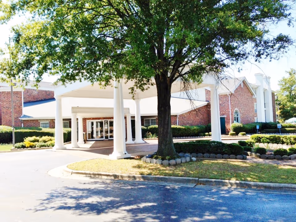 Exterior view of a senior living facility with a covered entrance supported by white columns, a large tree in the foreground, and well-maintained landscaping including bushes and grass around the building.