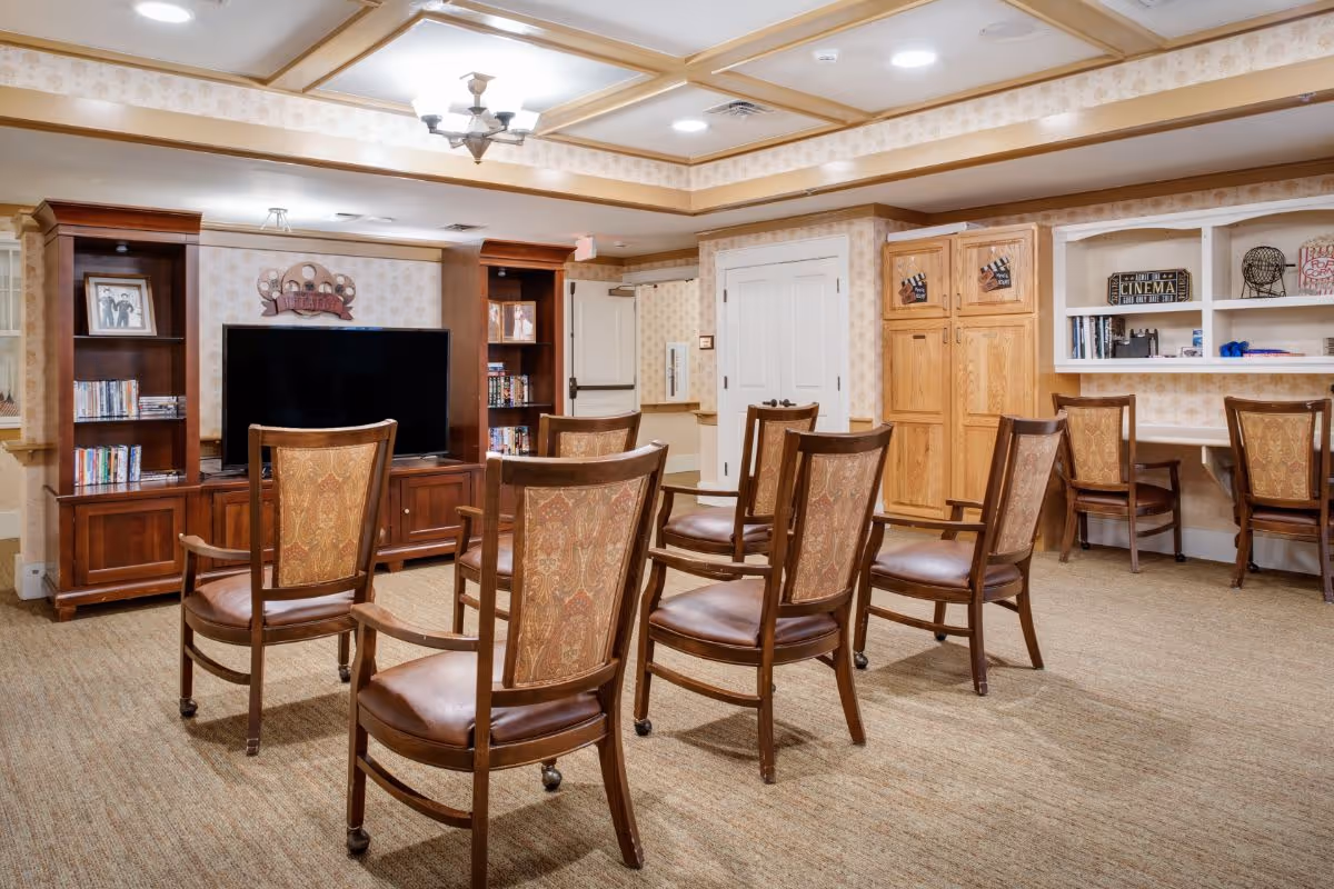 A cozy common area with eight wooden chairs arranged in two rows facing a large flat-screen TV on a wooden entertainment center. The room has beige patterned wallpaper, carpeted floor, and a ceiling with recessed lighting and a chandelier. There are bookshelves with DVDs and decorative items, and a built-in desk area with chairs on the right side.