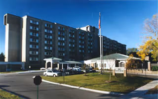 Exterior front view of a tall, dark-brick senior living building with a driveway, white entrance canopy, parked cars, and an American flag.