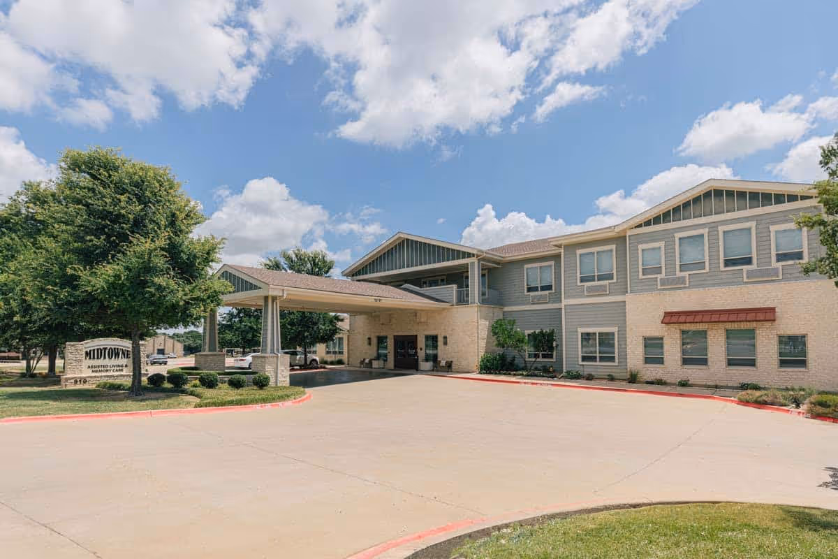 Exterior view of Midtowne Assisted Living and Memory Care building under a partly cloudy sky. The building is two stories with a covered entrance driveway, surrounded by trees and landscaping.