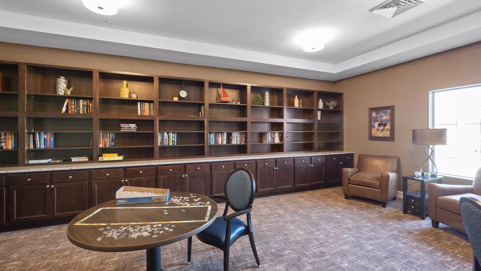 A spacious room with a large wooden bookshelf filled with books and decorative items along one wall. In the center, there is a round table with a partially completed jigsaw puzzle and a chair. Two brown leather armchairs are placed near a window with natural light, next to a small side table with a lamp. The room has a carpeted floor and neutral-colored walls.
