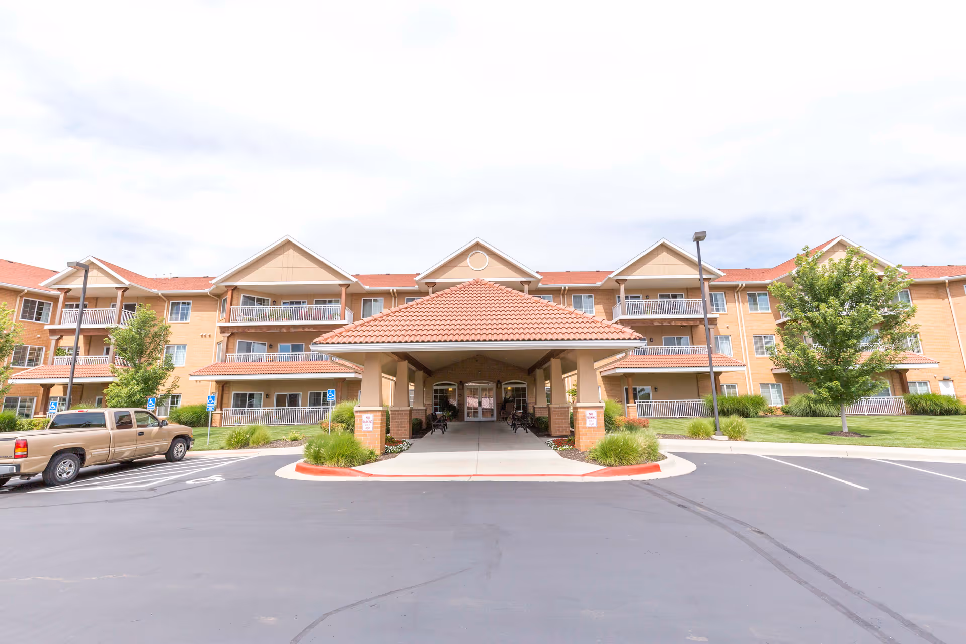 Front entrance of a three-story senior living building with a covered porte-cochere, balconies, and a parking lot.