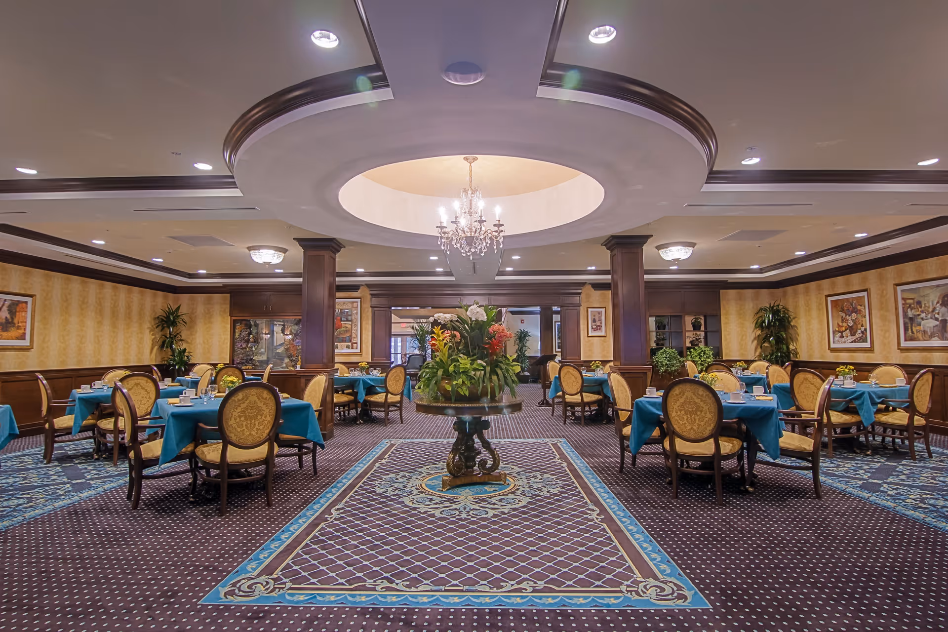 Elegant dining room with multiple round tables covered with teal tablecloths and set with cups and glasses. The room features ornate carpeting, yellow patterned wallpaper, wooden paneling, framed artwork on the walls, and a central chandelier hanging from a recessed ceiling. A large floral arrangement is placed on a round table in the center of the room.