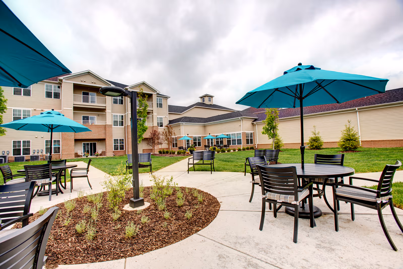 Outdoor patio area at StoryPoint Chesterton featuring multiple round tables with black chairs and teal umbrellas. The patio is surrounded by a well-maintained lawn and landscaping with small plants and trees. In the background, there is a multi-story beige building with balconies and large windows under a cloudy sky.