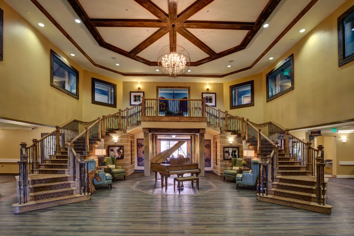 Spacious and elegant interior of a senior living facility featuring a grand double staircase with carpeted steps and wooden railings. In the center, there is a grand piano with a matching bench on a polished wooden floor. The ceiling has wooden beams arranged in a geometric pattern with a chandelier hanging in the middle. Comfortable armchairs are placed near the staircases, and framed artwork adorns the walls. Multiple windows allow natural light to enter the space.