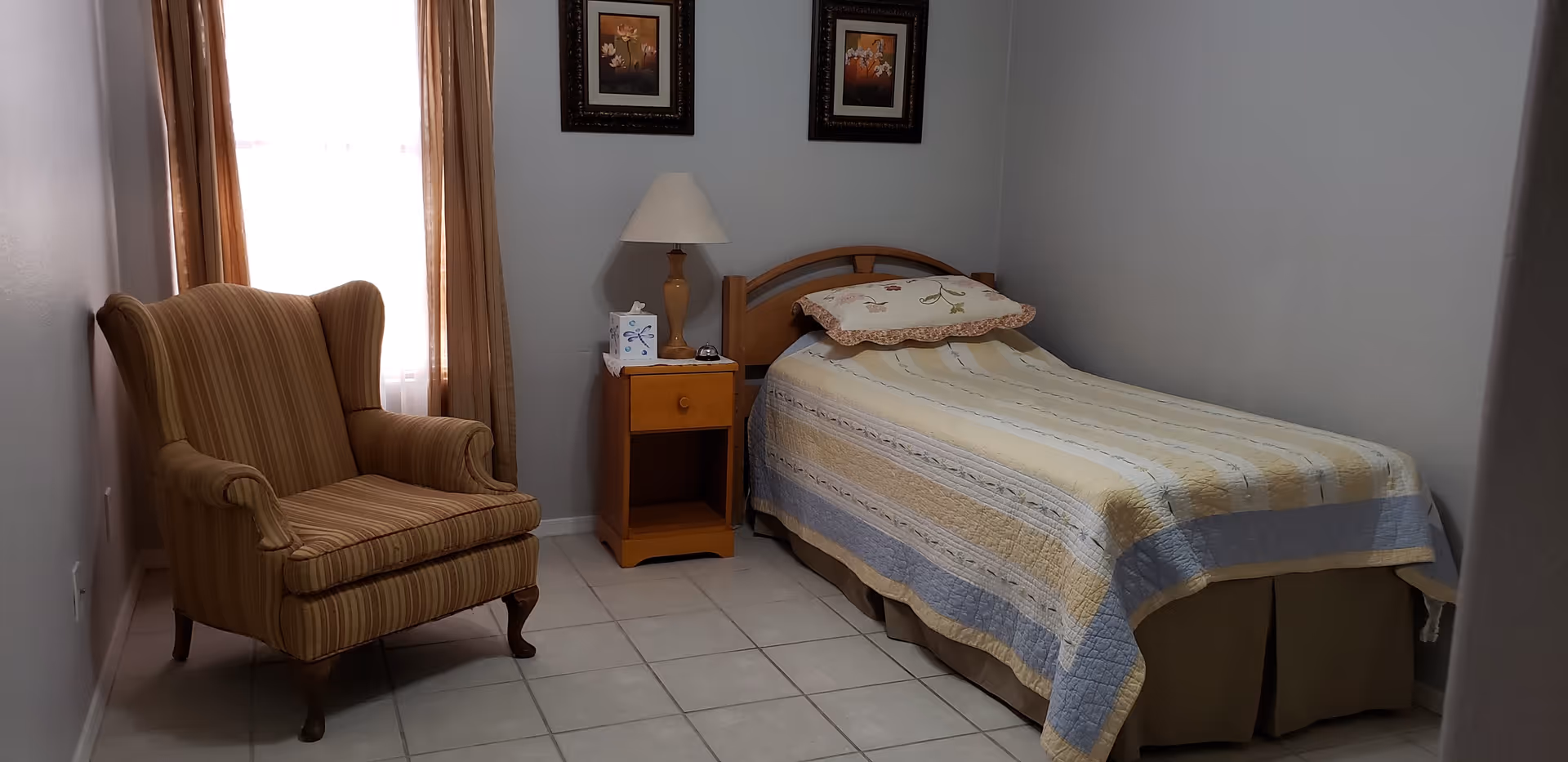Small bedroom with a single bed, nightstand and lamp, and a striped armchair by a curtained window.