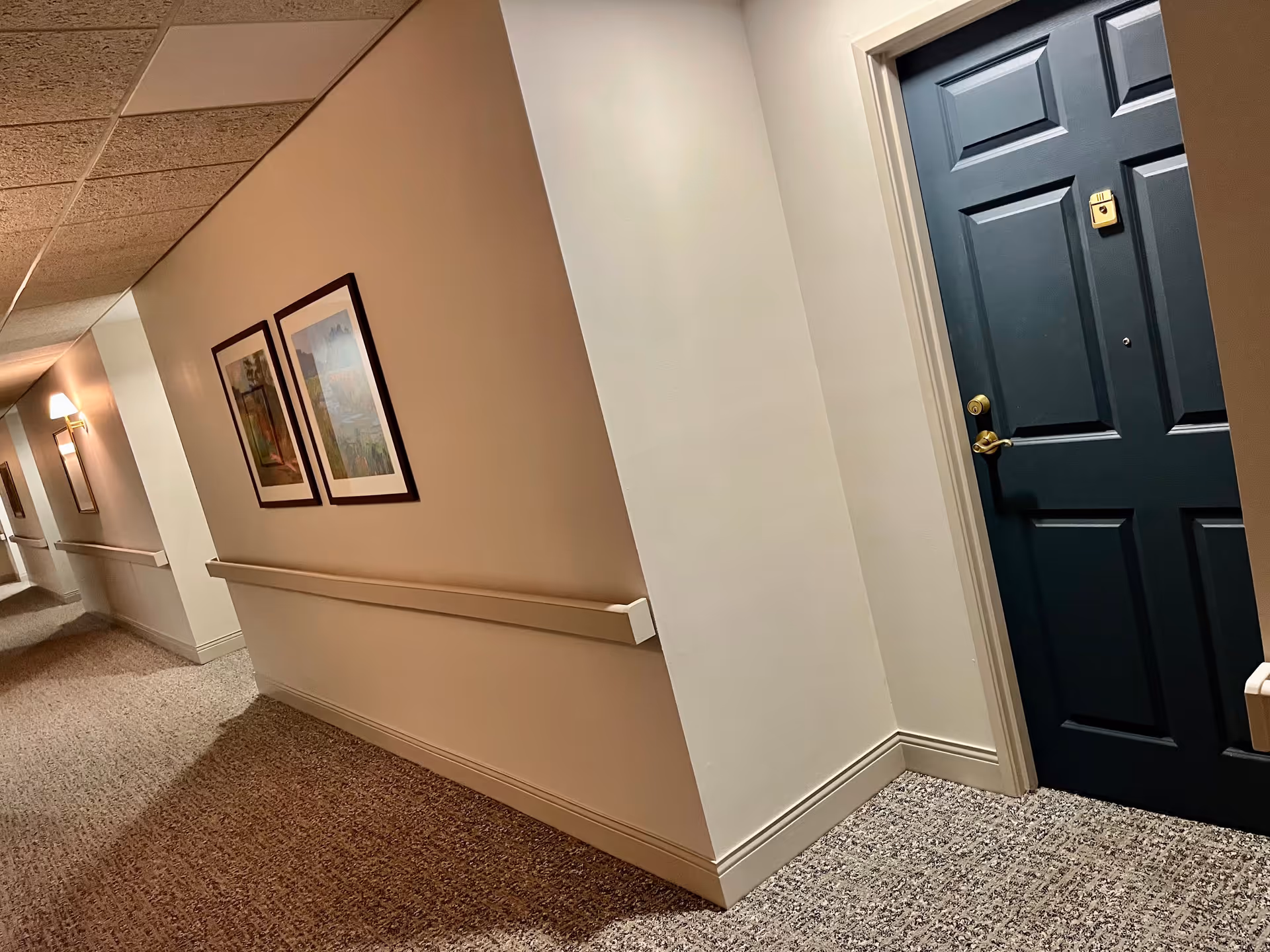 Interior hallway of a senior living facility with beige walls, carpeted floor, handrails along the walls, framed artwork, and a dark blue door with a gold handle and lock.