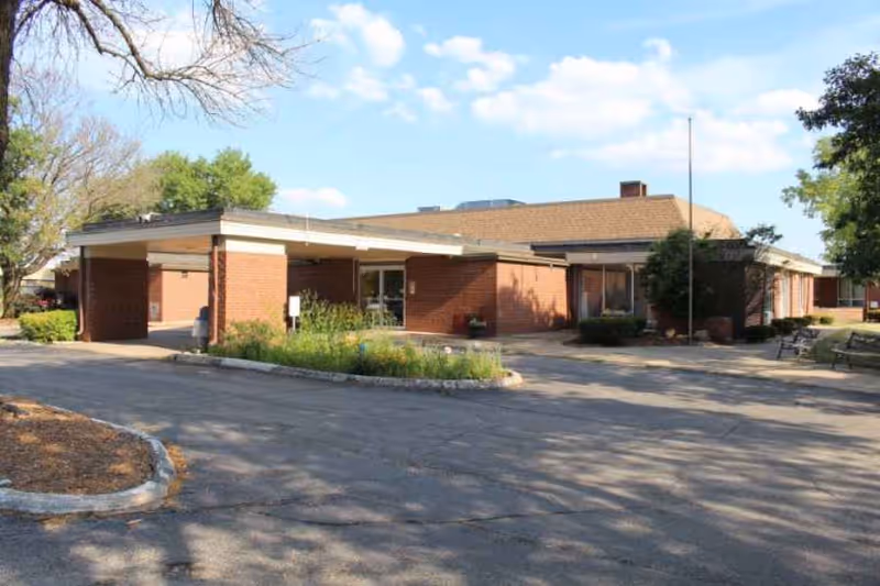 Single-story brick nursing home building with a covered entrance, driveway, and surrounding trees under a blue sky.