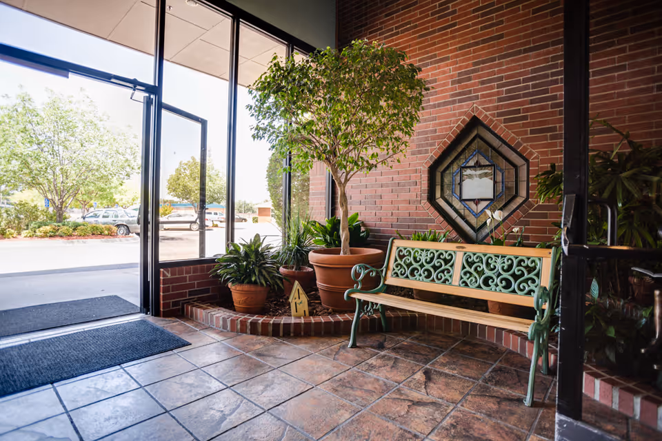 Sunlit entry lobby with potted plants and a decorative metal-and-wood bench against a brick wall near glass doors.