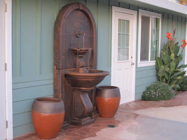 Outdoor area with a decorative wall-mounted water fountain flanked by two large brown planters, next to a white door and window on a light green building wall, with some plants and flowers nearby.