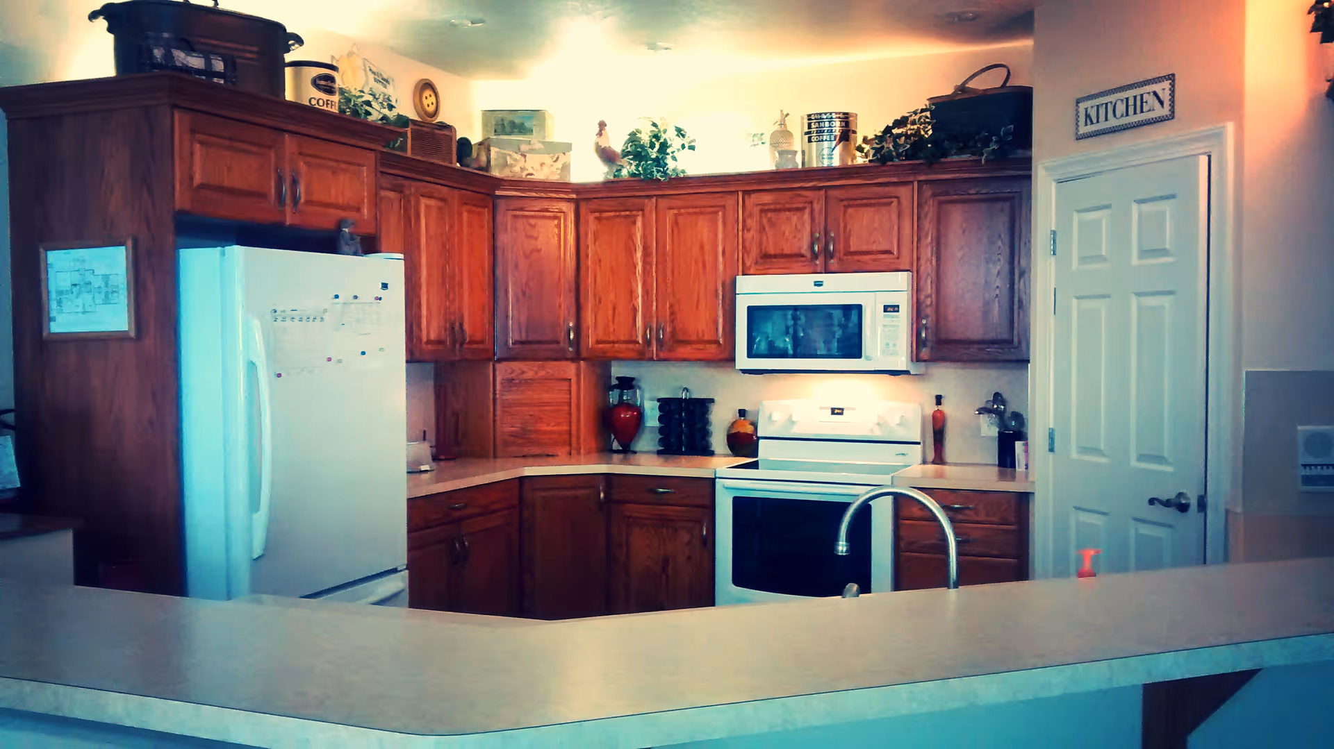 A kitchen area with wooden cabinets, a white refrigerator, a white microwave above a white stove, and a countertop with a sink and faucet. Decorative items and plants are placed on top of the cabinets. A door with a sign labeled 'KITCHEN' is visible on the right side.