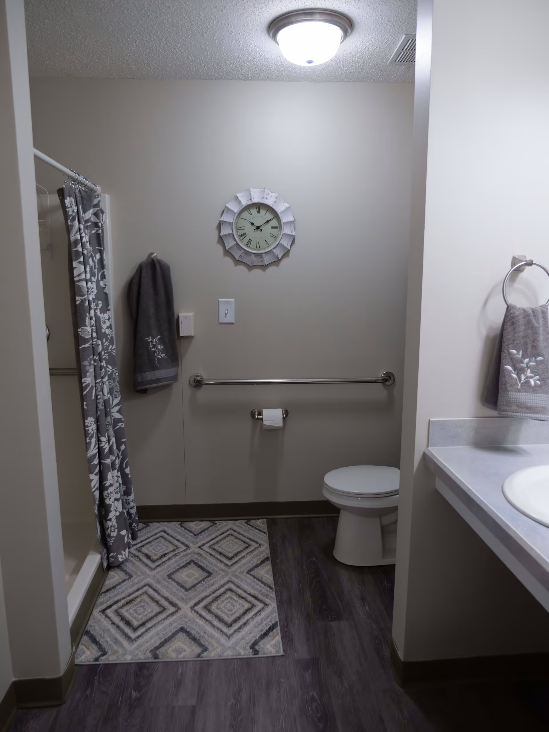 A bathroom with a shower area on the left featuring a floral patterned shower curtain. A gray towel hangs on a hook next to the shower. On the back wall, there is a decorative clock above a grab bar and a toilet paper holder with a roll of toilet paper. To the right, a white toilet is visible next to a countertop with a sink and another gray towel hanging on a ring. The floor has a dark wood-like finish with a patterned rug in front of the shower.