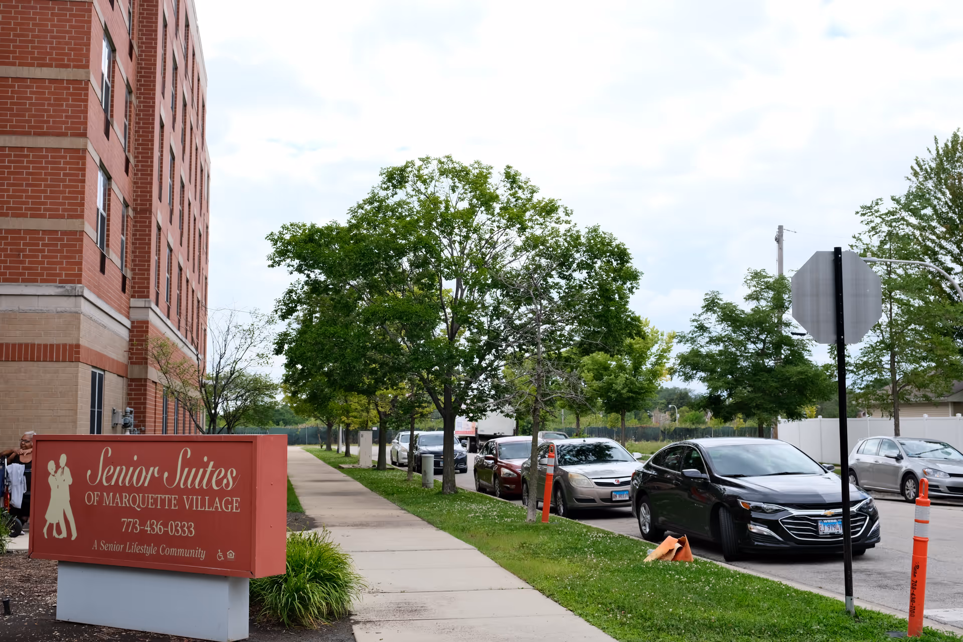 Sidewalk and entrance area of a brick senior living building with a 'Senior Suites of Marquette Village' sign, trees, and parked cars.