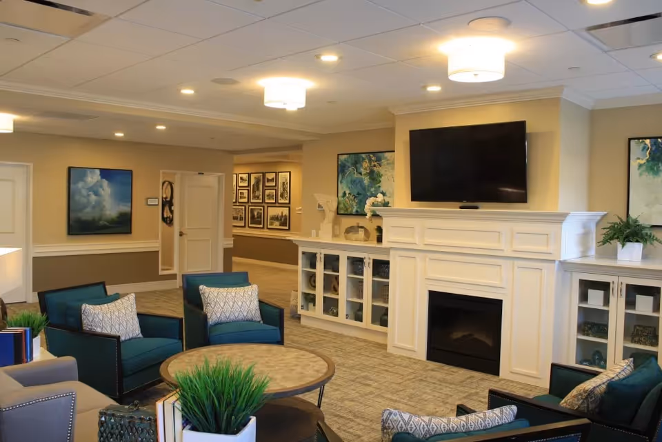 A cozy living room area in a senior living facility with blue armchairs, a round coffee table, a fireplace with a mounted flat-screen TV above it, decorative plants, and framed artwork on the walls.