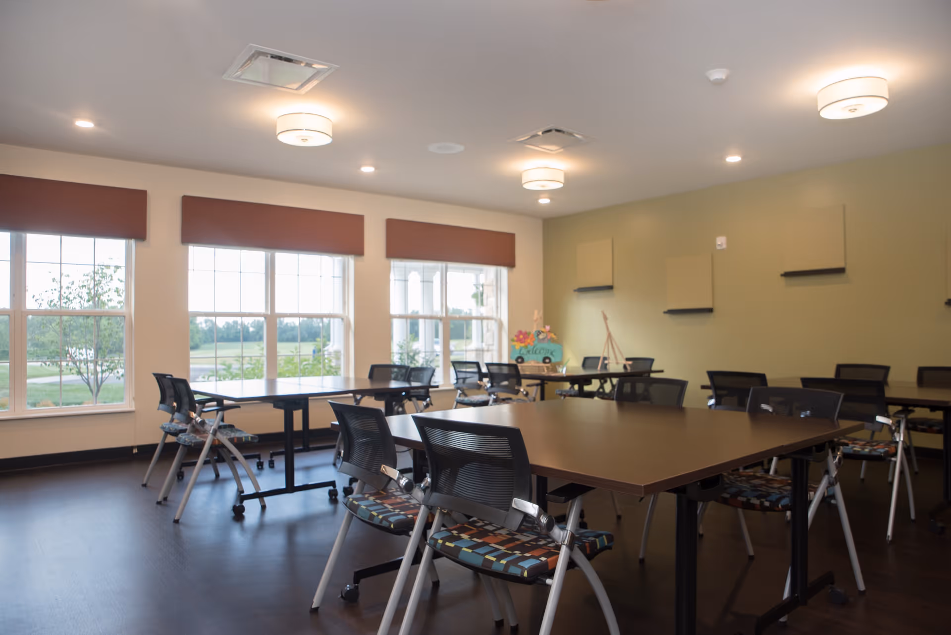 A bright and spacious room with multiple tables and chairs arranged neatly, large windows with brown valances letting in natural light, and a green accent wall with decorative panels and a welcome sign on an easel.