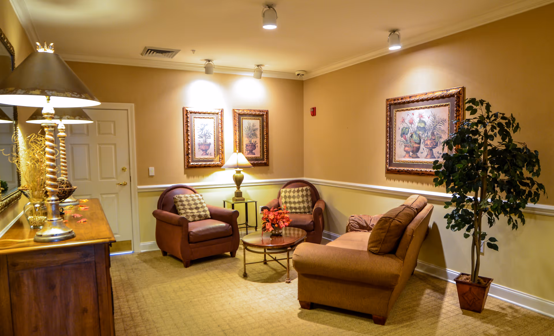A cozy sitting area in a senior living facility featuring two armchairs and a sofa arranged around a round glass coffee table with a floral centerpiece. The walls are painted beige with white trim and decorated with framed botanical artwork. A potted plant stands in the corner, and a wooden sideboard with a lamp and decorative items is visible on the left side.
