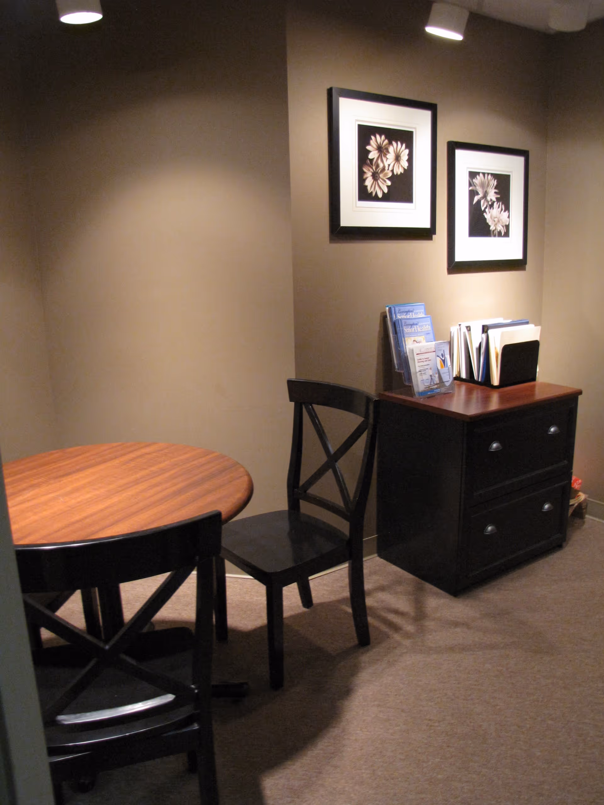 A small room with a round wooden table and two black chairs. There is a black cabinet with two drawers against the wall, holding brochures and file folders. Two framed floral pictures hang on the beige wall above the cabinet.