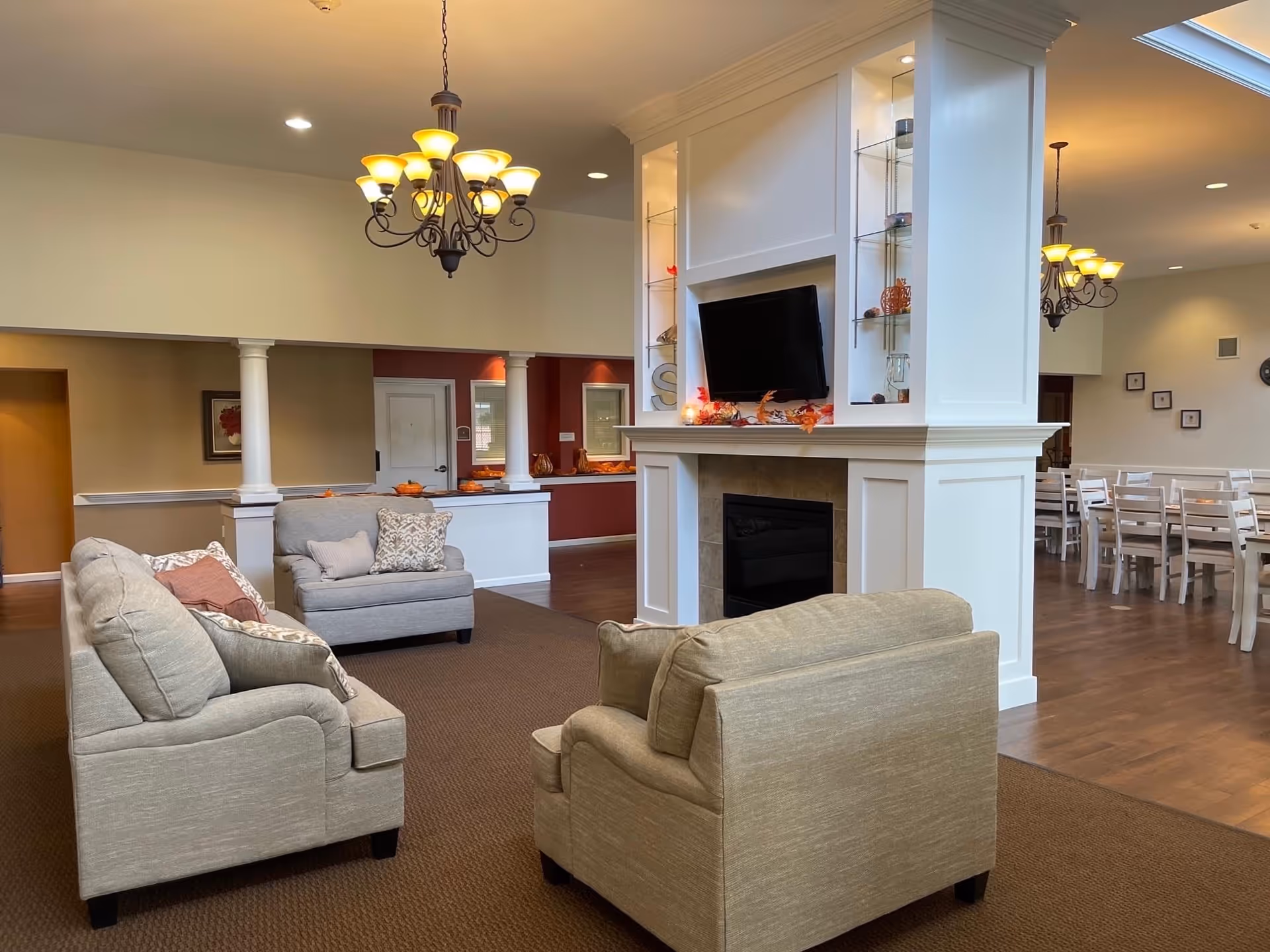 A cozy living room area with beige upholstered sofas and armchairs arranged around a white fireplace with a mounted flat-screen TV. The fireplace has built-in glass shelves with decorative items. In the background, there is a dining area with white tables and chairs, wooden flooring, and warm lighting from chandeliers.