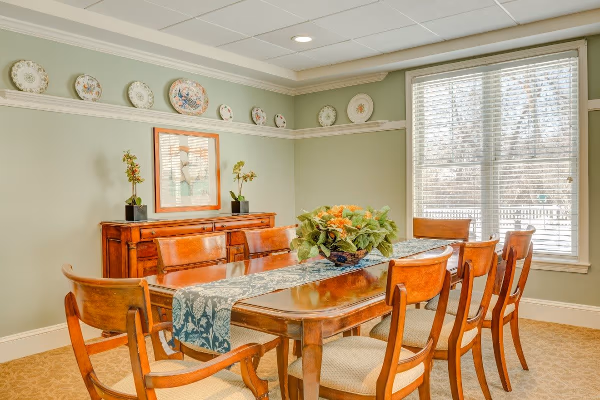 A dining room with a wooden dining table and six matching chairs. The table has a blue patterned runner and a centerpiece with green and orange flowers. Behind the table is a wooden sideboard with two small potted plants and a framed picture above it. Decorative plates are displayed on a shelf running along the upper part of the light green walls. A large window with white blinds lets in natural light.