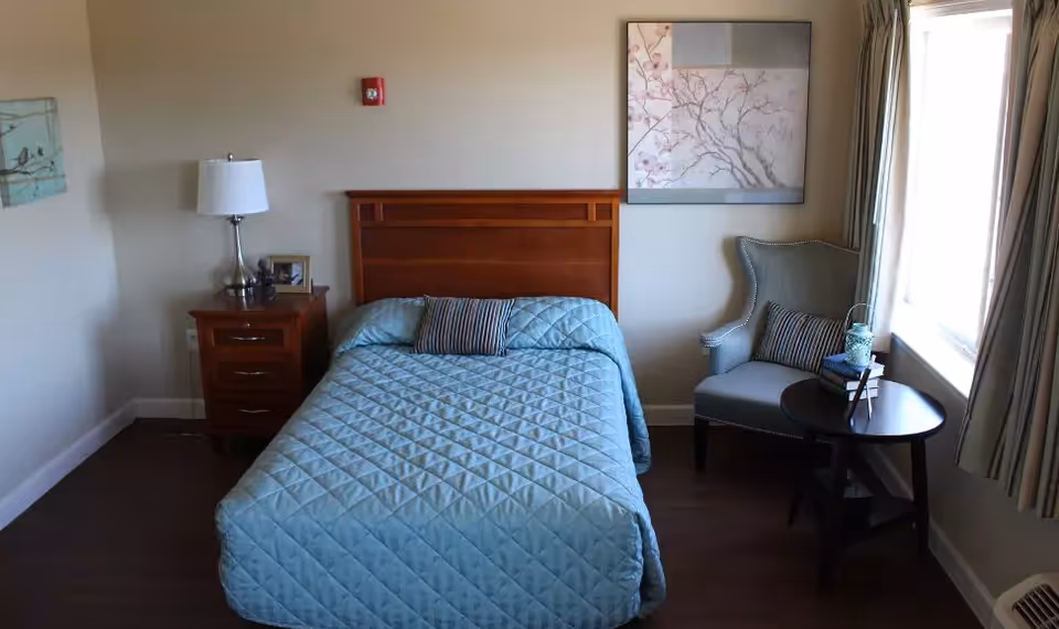 A neatly made bed with a light blue quilt and a striped pillow in a senior living facility room. To the left of the bed is a wooden nightstand with a lamp and a small picture frame. On the right side, there is a comfortable armchair with a striped pillow and a round wooden side table holding a decorative lantern and some books. A large window with curtains allows natural light to brighten the room. The walls are decorated with framed artwork.
