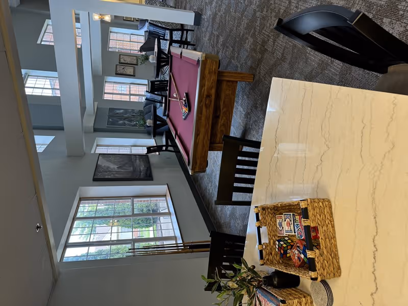 Interior view of a recreational common area in a senior living facility with a pool table in the center, several tables and chairs near large windows letting in natural light, and a countertop with baskets containing playing cards and a Rubik's cube.