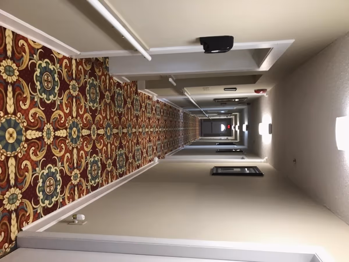 A long indoor hallway with beige walls and a colorful patterned carpet featuring red, blue, and yellow floral designs. The hallway is lined with doors on both sides and illuminated by wall-mounted lights. There is a fire alarm and a security camera visible on the ceiling.