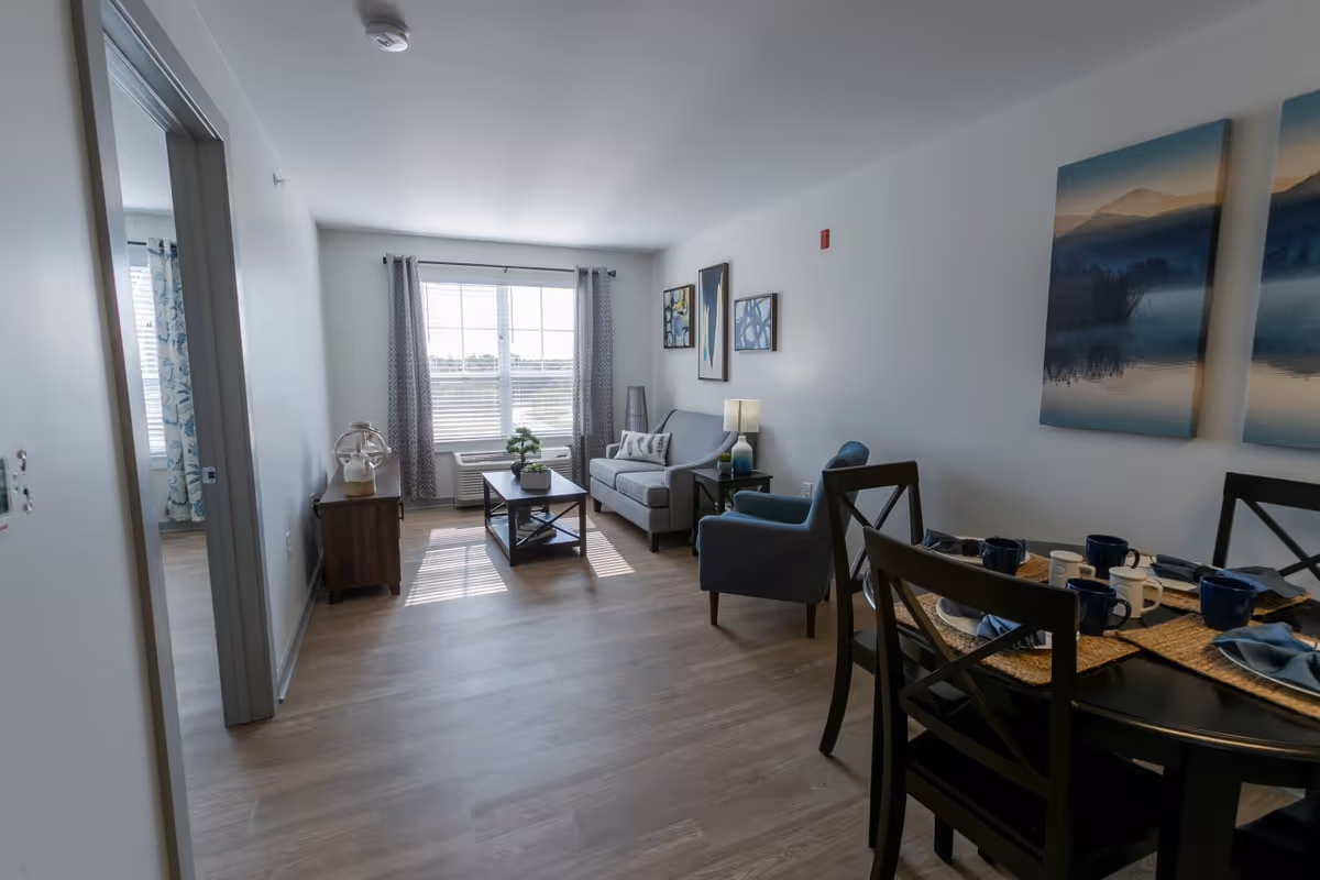 Sunny open living area with a sofa, armchairs, coffee table and a dining table set beside a large window.
