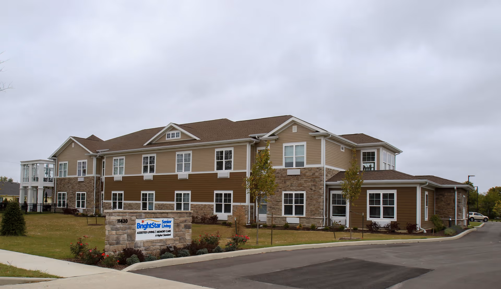 Two-story assisted living building with stone and beige siding, landscaped lawn, driveway and a stone sign in front under a gray sky.