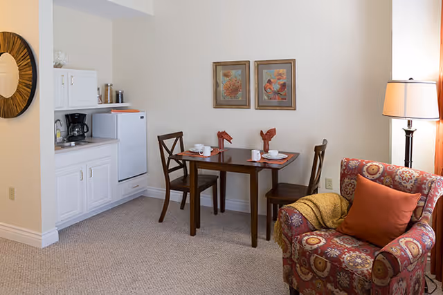 A cozy senior living area featuring a small kitchenette with white cabinets, a coffee maker, and a mini fridge. Adjacent to the kitchenette is a wooden dining table set for two with cups and napkins, flanked by two wooden chairs. A patterned armchair with an orange pillow and a yellow throw blanket sits next to a floor lamp with a white shade. Two framed floral artworks hang on the beige wall above the dining table.