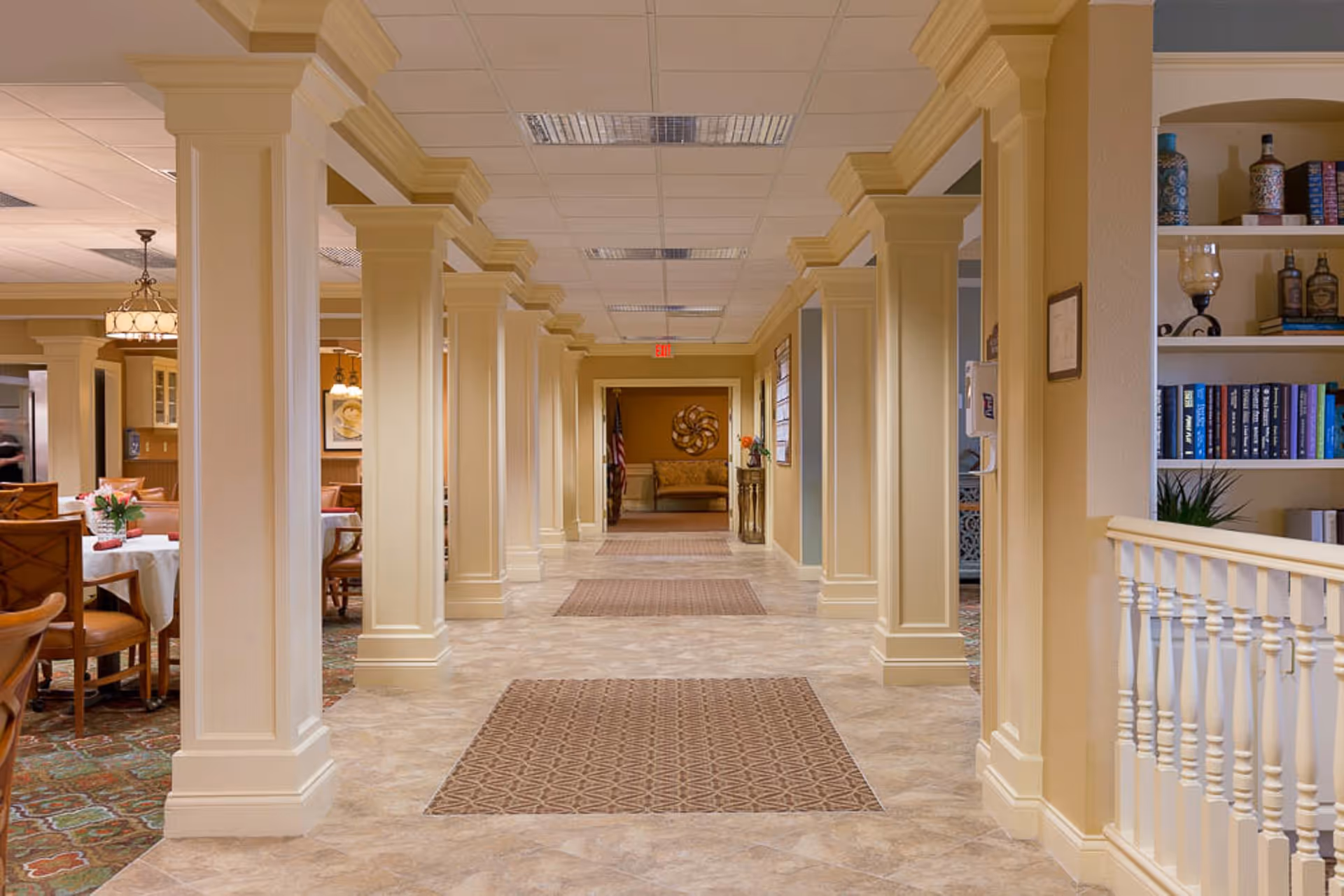 Interior hallway of The Bridge at Paradise Valley featuring cream-colored columns, beige tiled floor with patterned rugs, and a dining area with tables and chairs to the left. Shelves with books and decorative items are visible on the right side.