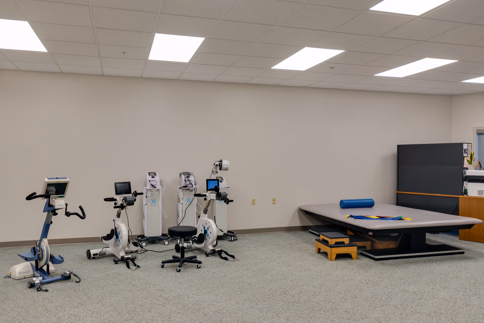 A therapy or exercise room with stationary exercise bikes, medical equipment, and a large padded therapy table with a blue foam roller and colorful resistance bands on it. The room has beige walls, a carpeted floor, and bright overhead lighting.
