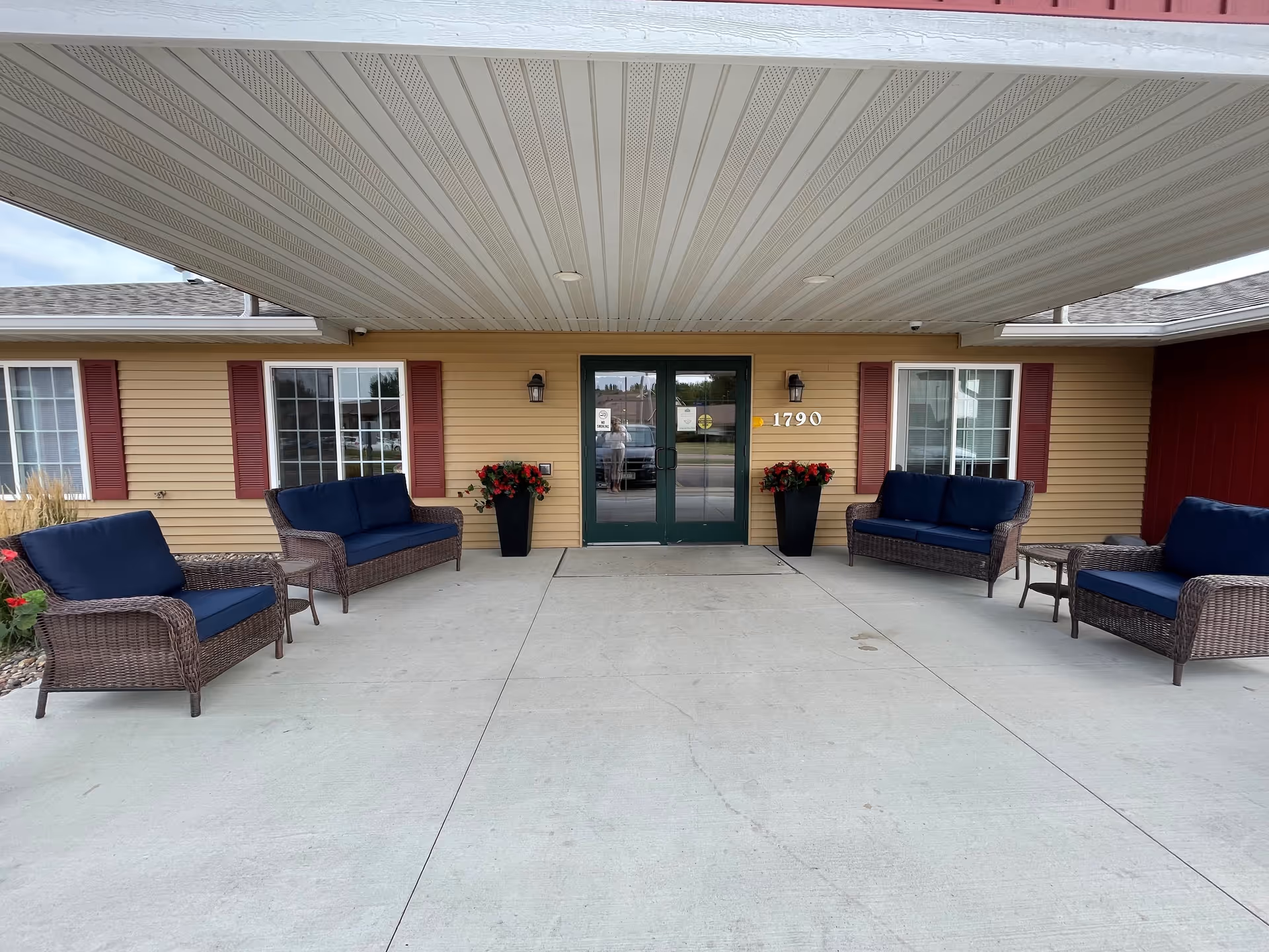 Entrance of a senior living facility with a covered porch area featuring four wicker chairs with blue cushions and two small tables. The building has beige siding with red shutters on the windows and two black planters with red flowers flanking the green double doors. The address number 1790 is displayed next to the door.
