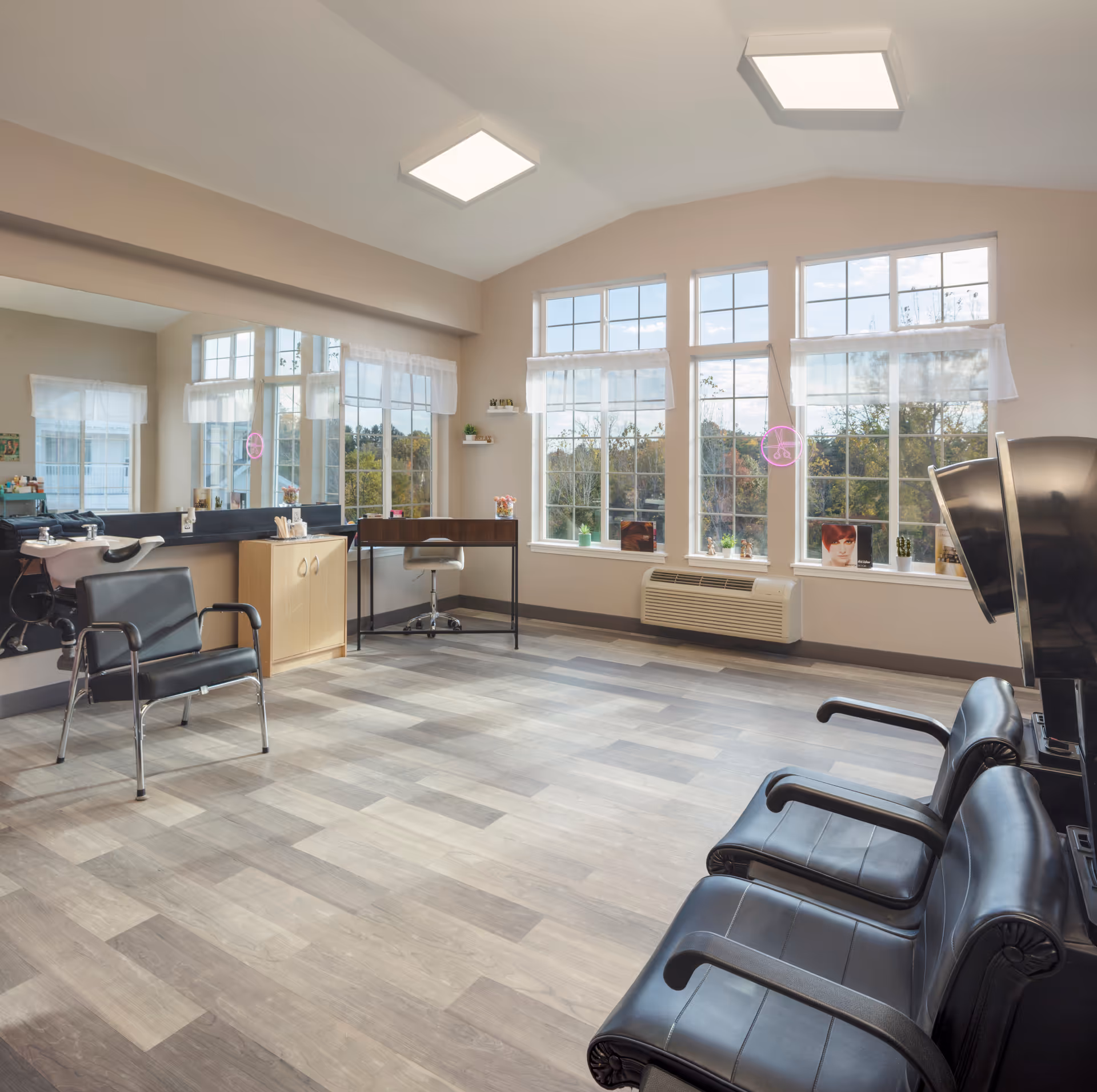 A bright and spacious hair salon area with large windows letting in natural light. The room features black salon chairs, a hair washing station, a small desk with a chair, and a large mirror along one wall. The floor is covered with light wood-patterned vinyl, and there are small plants and hair product displays on the windowsills.