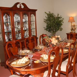 Formal dining room with a polished wooden table set with plates, glassware and a floral centerpiece, matching chairs, a china cabinet and lamp.