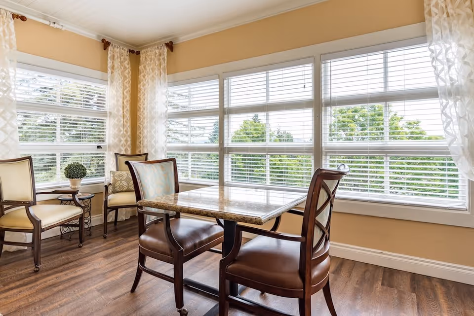 A bright corner room with large windows covered by white blinds and sheer curtains. The room features a square marble-top table with two wooden chairs with leather seats, and two additional upholstered chairs with wooden frames near a small round side table holding a potted plant. The floor is wooden and the walls are painted beige.