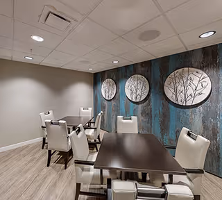 Interior view of a dining area with two dark wooden tables and white cushioned chairs. The room has light wood flooring, a white ceiling with recessed lighting, and a wall decorated with three circular art pieces featuring botanical designs on a textured blue and brown background.
