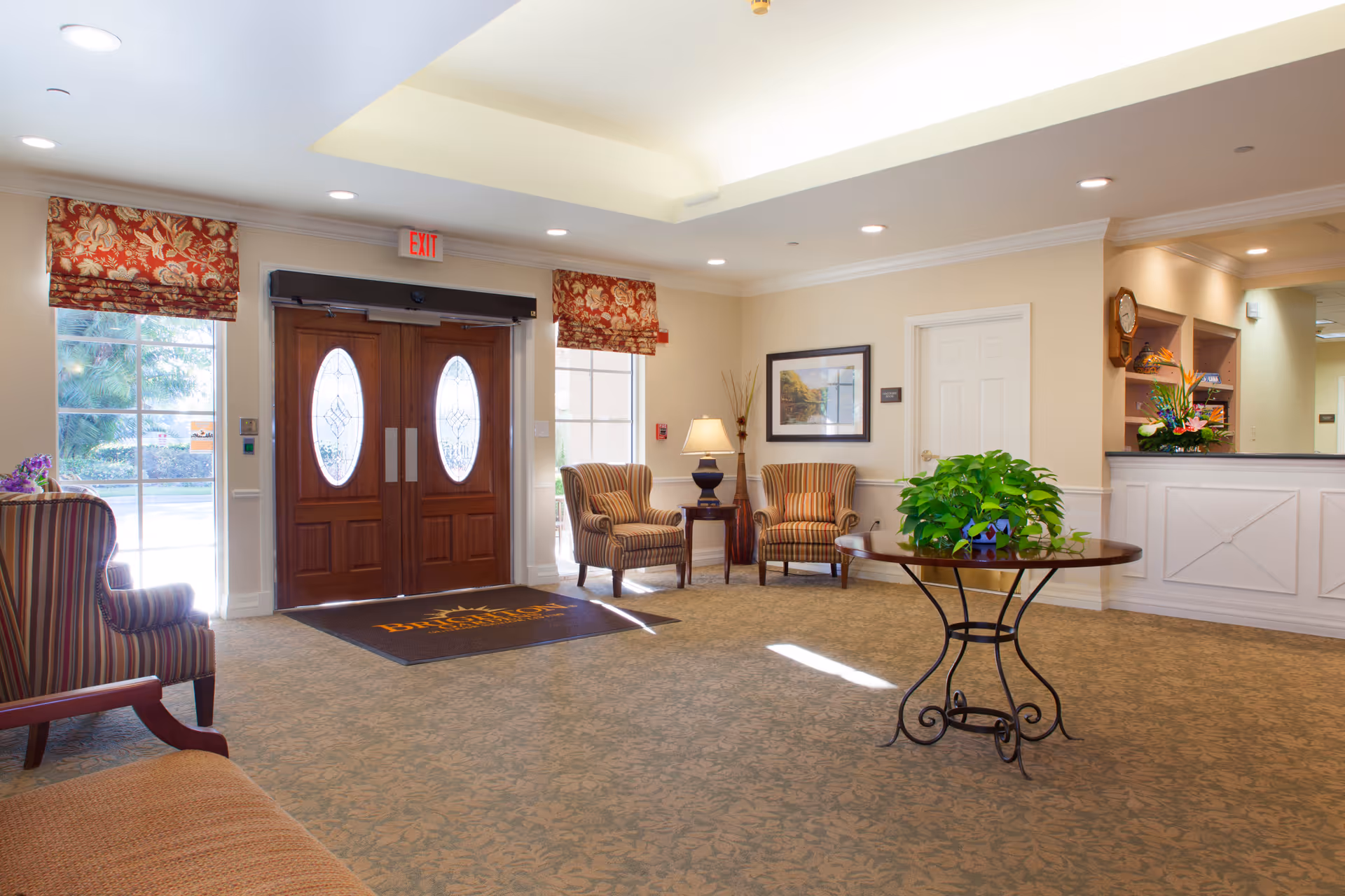 Bright senior living facility lobby with double wooden entrance doors, seating area, a reception desk, and a round table holding a potted plant.