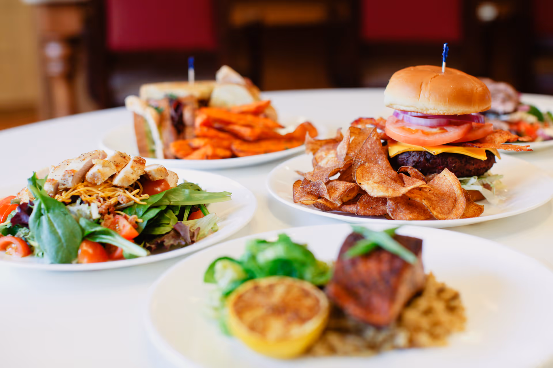 A close-up view of a table with several plates of food including a chicken salad, a cheeseburger with potato chips, a sandwich with sweet potato fries, and a dish with grilled fish, lemon, and vegetables.