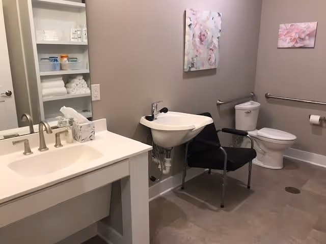 A clean and accessible bathroom with a white sink and countertop on the left, a small wall-mounted sink with a black chair in front of it, a toilet with grab bars on the right wall, and two floral paintings hanging on the beige walls. Shelves with towels and toiletries are visible in the mirror reflection.